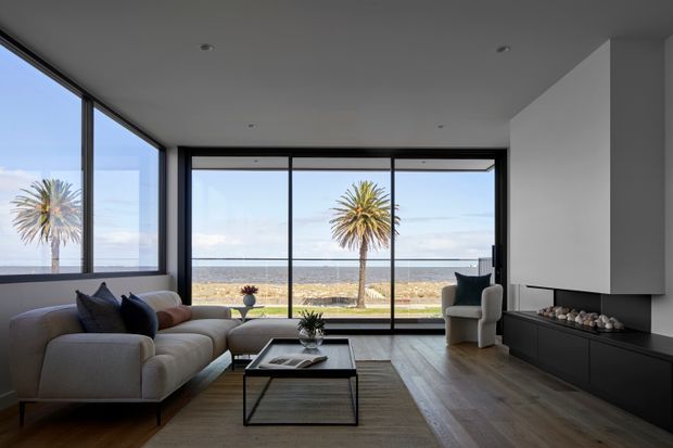 Contemporary living room with a large sectional sofa, low rectangular coffee table, sculptural armchair, and streamlined dark built-in media cabinetry. Floor-to-ceiling black-framed glazing provides expansive views of a beach, ocean, and palm trees. The room uses a restrained neutral palette, recessed ceiling lighting, and warm timber flooring.