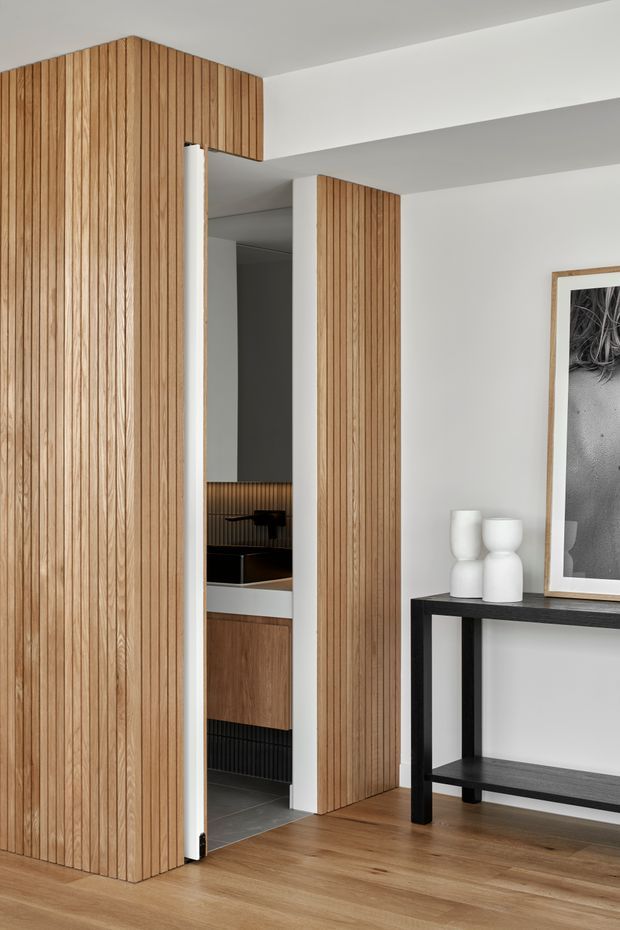 A refined contemporary interior showing a timber-lined concealed doorway and matching vertical timber wall panelling. The warm wood flooring and panelling contrast with crisp white walls and ceiling planes. Through the partially open door, a compact bathroom or powder room is visible with dark tiled flooring, timber vanity cabinetry, a black basin, and dark vertical wall detailing. A black console table with white sculptural vessels and framed monochrome artwork completes the entry space.