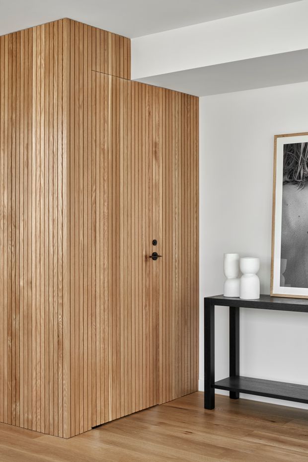 A minimalist interior entry area featuring a full-height timber door and adjoining wall clad in narrow vertical timber battens. The warm wood continues onto the floor, contrasted with white walls and ceiling beams. A slim black console table with white sculptural vessels and a framed monochrome artwork sits to the right. The composition is clean, restrained, and contemporary.