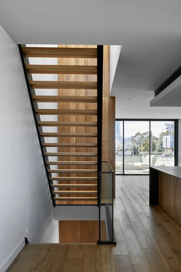 A contemporary interior view focused on an open-riser timber staircase with dark metal stringers and glass balustrading. The staircase connects a timber-floored landing to an open-plan living area with full-height glazing, timber wall panelling, and built-in timber cabinetry. The space is bright and minimalist, with strong linear architectural detailing.