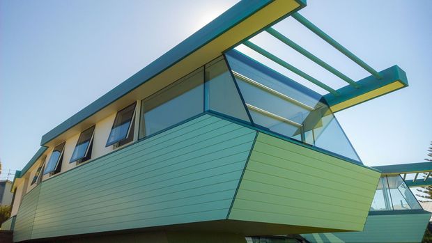Exterior view of a distinctive modern, angular multi-level building with extensive turquoise-green horizontal cladding, dark window frames, large glazed openings, and a projecting roof canopy with exposed green structural beams. The design has a strong geometric, cantilevered form and a bright open-air architectural character.