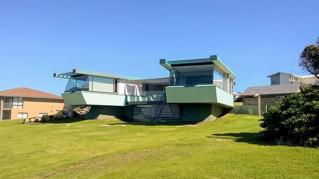 Exterior view of a distinctive elevated mid-century-style house set within a broad manicured lawn. The building has low-pitched roof forms, projecting geometric volumes, horizontal green cladding, extensive glazing, open balcony or terrace areas, and an external stair leading to the raised main level. Neighbouring suburban houses and landscaping are visible in the background under a clear blue sky.