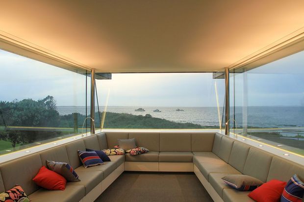 A spacious enclosed coastal lounge with a large U-shaped sectional sofa arranged around the room. Full-height corner glazing provides panoramic views over dense coastal vegetation and the ocean, while a broad recessed ceiling and warm integrated lighting create an inviting atmosphere. The interior uses neutral upholstery, dark flooring, and colourful cushions as accents.