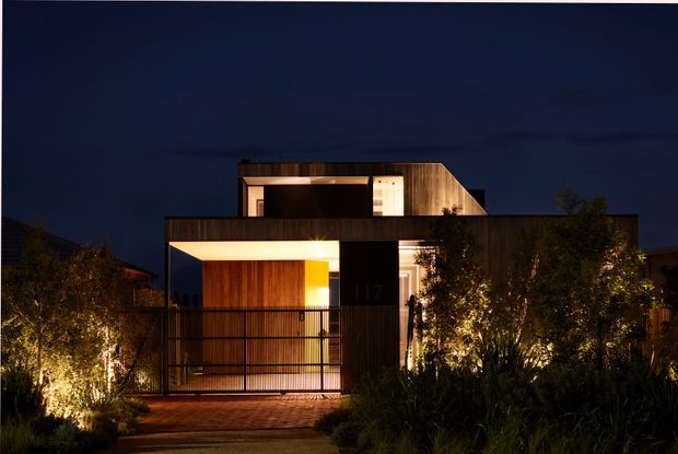 Night-time photograph of a contemporary multi-level residential facade. The dark geometric building features warm timber cladding, a projecting flat-roofed entry canopy, illuminated glazing, black metal fencing, and carefully lit front landscaping.