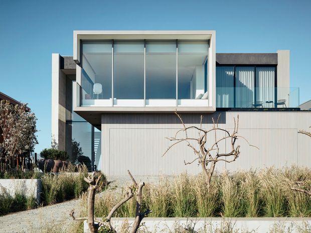 Exterior view of a contemporary multi-level residence with strong rectilinear forms, extensive floor-to-ceiling glazing, a projecting upper-level glazed volume, dark structural elements, and pale vertically ribbed cladding. The landscaped foreground includes ornamental grasses and pruned deciduous trees.