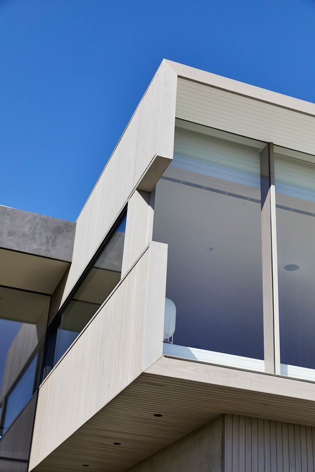 Upward-looking close view of a contemporary multi-level building facade beneath a clear blue sky. The exterior combines pale vertically textured cladding, dark-framed glazing, projecting balcony or cantilevered volumes, and a recessed soffit lined with narrow horizontal boards and recessed downlights. A large glazed opening reveals a bright interior area, while a grey rendered wall forms part of the adjoining structure.