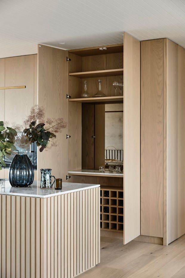 A warm, refined kitchen or dining entertaining area featuring extensive light timber cabinetry, a concealed bar and wine-storage joinery, open shelves displaying glassware, and a central island with a vertically fluted timber front and pale stone benchtop. The space has light timber flooring, a white linear-panel ceiling, and restrained neutral styling with decorative glassware and foliage.