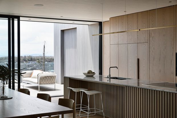 A refined contemporary open-plan kitchen and dining area with a large timber island, integrated timber cabinetry, dark stone benchtop, and vertical timber slat detailing. Floor-to-ceiling black-framed glazing opens to a bright outdoor terrace and distant landscape. A timber dining table, bar stools, pendant lighting, and a sculptural indoor plant complete the space.