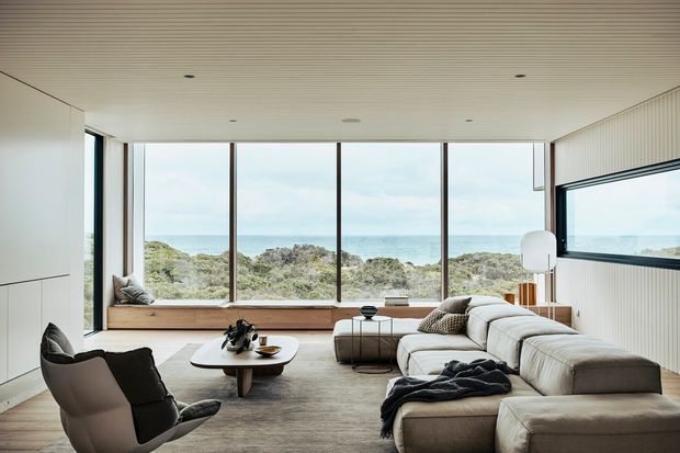 A bright, contemporary living room with a large L-shaped sectional sofa, armchairs, low coffee table and floor lamp. Full-height glazing frames a coastal ocean view, while a built-in window seat runs along the glazed wall. The room has a pale neutral palette, a finely linear ceiling, vertical wall panelling, and dark slender window frames.