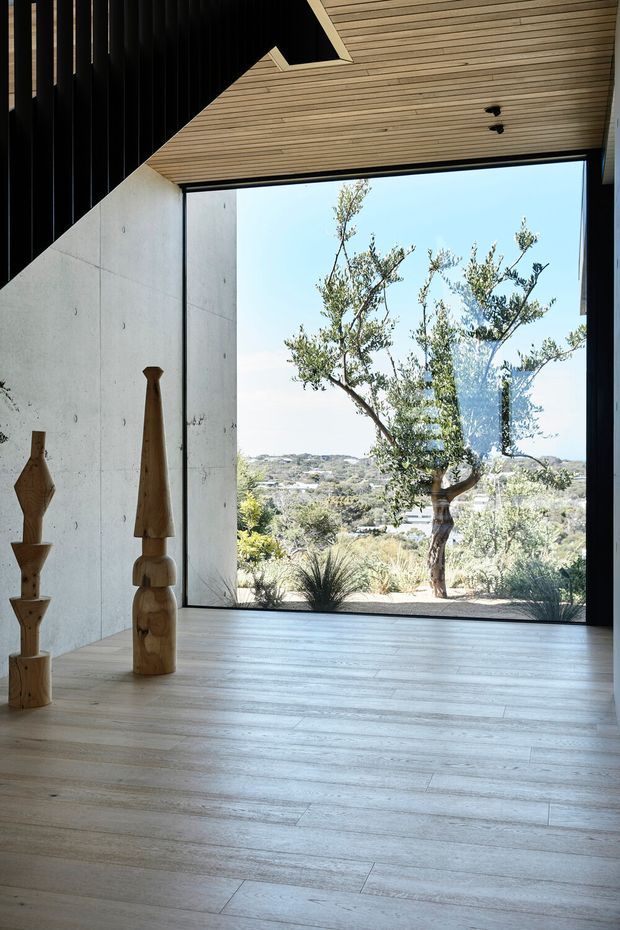 Minimalist interior with a large frameless glazed opening overlooking a landscaped, hilly suburban setting. The room features pale timber-look flooring, a fully exposed smooth concrete wall, a slatted timber ceiling, black vertical timber or metal detailing, and sculptural wooden objects. The space is bright, airy, and strongly connected to the landscape.