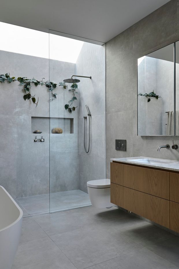 Contemporary minimalist bathroom with a walk-in glass shower, freestanding bathtub partially visible at left, wall-hung timber vanity, integrated basin, large mirror, wall-mounted fixtures, and grey concrete-look tiled surfaces. A small indoor trailing plant adds an organic accent.