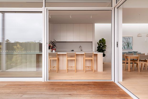 A bright, contemporary open-plan kitchen and dining area viewed from a timber deck through wide glazed sliding or folding doors. The kitchen features pale wood cabinetry, a long island with a light benchtop and integrated seating, white walls, and minimal pendant-free ceiling lighting. A timber dining table and chairs are visible to the right, with indoor plants and artwork adding warmth. The extensive glazing connects the interior to the deck and landscaped outlook.