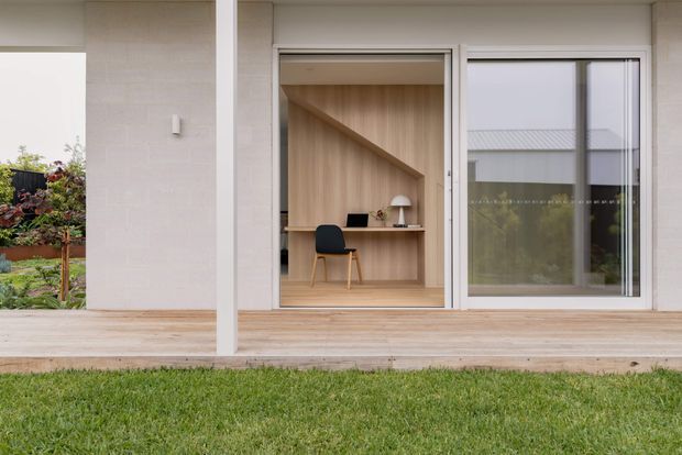 Exterior view of a contemporary single-storey home showing a covered timber deck, pale masonry facade, large glazed openings, and a compact built-in home office nook visible through the central sliding doors. The office features warm timber wall lining, an integrated desk, chair, and task lamp. A landscaped garden and lawn are visible around the building.