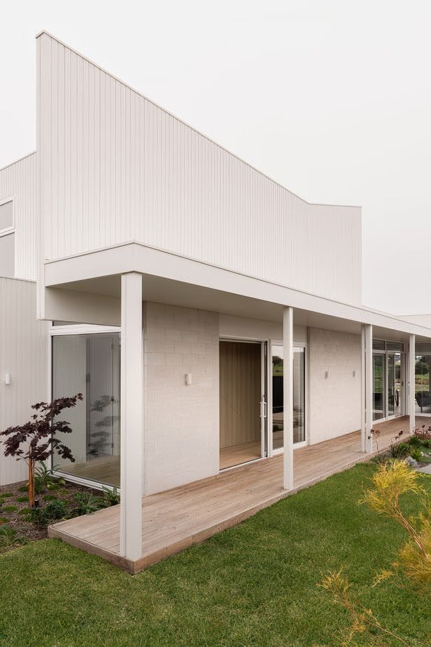 Exterior view of a contemporary single-storey building with a dramatic sloping vertical wall and pale ribbed cladding. A covered verandah runs along the facade, supported by slender white columns, with full-height glazed doors and windows opening onto a timber deck and landscaped lawn.