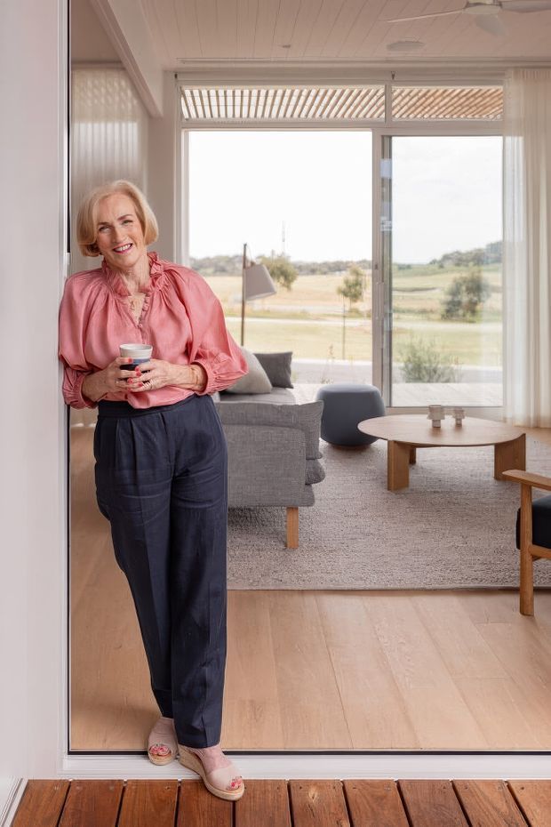 Bright contemporary living room viewed from a timber deck through a wide glazed opening. A woman stands in the foreground holding a cup, while the room contains a grey upholstered sofa, rounded dark lounge chair, round timber coffee table, and a timber-framed chair. Pale timber flooring, light neutral finishes, expansive glazing, sheer curtains, and a slatted overhead screen create an airy connection to the landscape beyond.