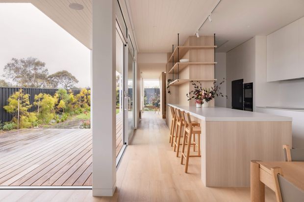 Bright open-plan kitchen and dining area opening onto a spacious timber deck and landscaped garden. The room features light timber flooring, pale timber cabinetry and open shelving, a large white island with timber stools, extensive glazing, and a clean contemporary fit-out. A dining table is partially visible in the foreground.
