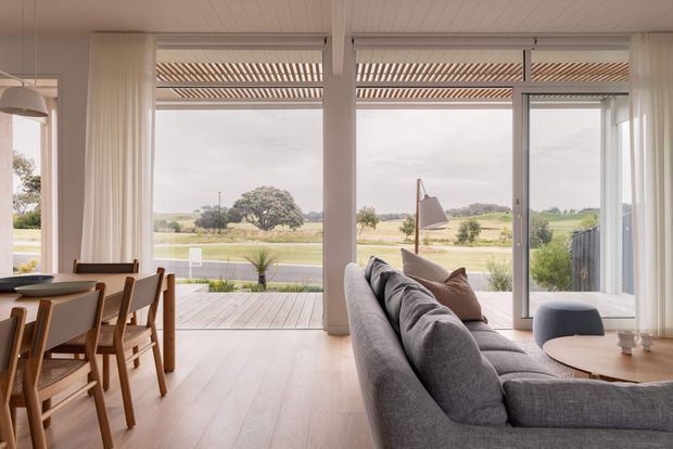A bright, open-plan living and dining area with floor-to-ceiling glazing overlooking a rural landscape. The room features pale timber flooring, a white-lined ceiling, white structural columns, sheer curtains, a grey sectional sofa, a round coffee table, and a timber dining table with woven-seat chairs. Exterior timber decking and a pergola-like overhead structure are visible beyond the glazing.