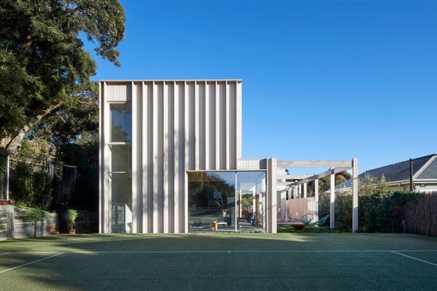 Exterior view of a contemporary multi-level residential extension with a tall rectangular volume clad in vertically ribbed light-toned panels and extensive glazing. A lower glazed living area opens toward a covered outdoor structure and a broad artificial-turf recreational court in the foreground. Mature trees and neighbouring residential buildings frame the suburban setting.