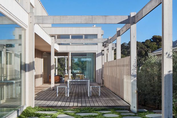 Contemporary multi-level residential exterior with a timber-clad deck and outdoor dining area framed by pale structural posts and beams. Large glazed doors and windows connect the deck to the interior, while horizontal clerestory glazing and clean rectilinear forms create a bright, open architectural composition. A timber privacy screen and stepping-stone landscape sit in the foreground.