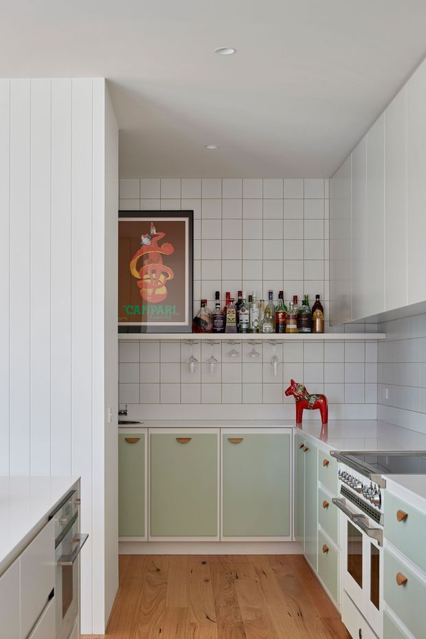 A compact, highly organized kitchen with pale mint-green lower cabinetry, white vertically grooved tall cabinetry, white tiled walls with dark grout, and warm timber flooring. A tiled splashback and open shelf form a small home bar area stocked with bottles and hanging stemware. Integrated stainless-steel appliances and a range cooker line the foreground, while a framed Campari poster and small red decorative object add colourful accents. The room is bright, clean, and retro-inspired.