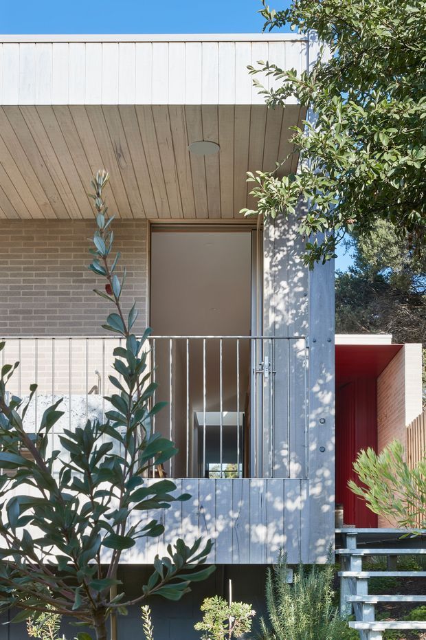 A close-up exterior view of a contemporary multi-level dwelling. The facade combines pale grey painted vertical cladding, warm grey brickwork, and a timber-lined recessed entry canopy. A metal balustrade fronts an upper-level doorway or balcony, while a vivid red recessed entrance volume and external stairway add contrast. Native-style planting partially screens the facade.