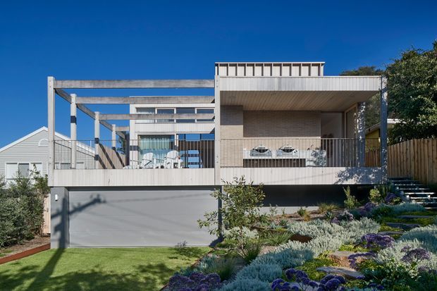 Front exterior view of a contemporary multi-level residence on a landscaped sloping site. The house features pale horizontal cladding, exposed structural framing, a broad covered upper terrace, large glazed openings, a rooftop privacy balustrade, and an integrated garage. Layered drought-tolerant planting and steps lead up beside the building.