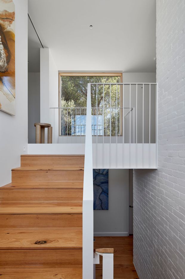 Interior view of a modern stairway with broad warm timber treads ascending beside a white-painted brick wall. A white metal balustrade overlooks the lower level, with a large timber-framed window providing natural light and views of trees. Minimal white walls, recessed ceiling lighting, and glimpses of artwork and lower-level flooring complete the composition.