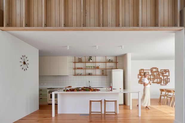 Bright open-plan kitchen and dining area with a white island, pale cabinetry, tiled splashback, timber flooring, open timber shelving, and a freestanding refrigerator. Vertical timber wall cladding forms a prominent upper-level or mezzanine feature, while a dining table and geometric wall artwork are visible beyond the kitchen.
