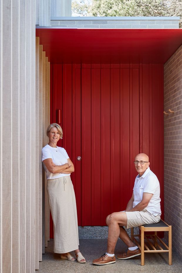 A portrait-oriented exterior architectural photograph showing two people posed at the entrance of a contemporary building. The entrance is framed by a vivid red vertically panelled door and matching red soffit, with pale vertical cladding on the left, exposed light-grey masonry above, and brown brickwork on the right. A narrow paved entry area and timber stool are visible.