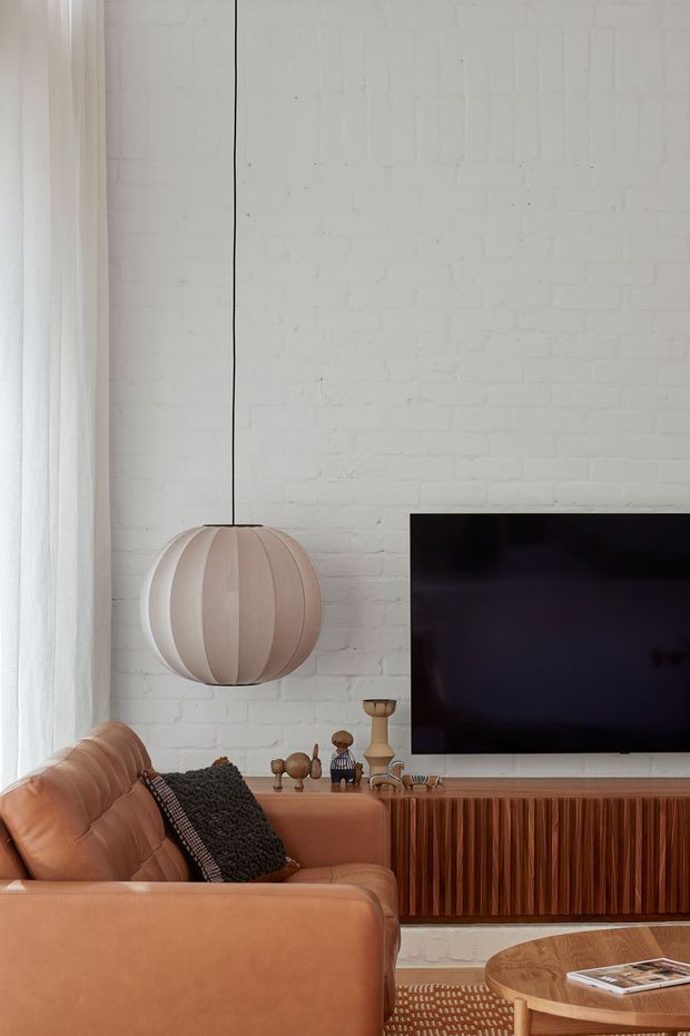 A warmly styled living room with a caramel leather sofa in the foreground, a textured dark cushion, a large pleated pendant light, and a wall-mounted television. The room features a white-painted exposed brick wall, sheer curtains, a fluted timber media console, decorative objects, and a round timber coffee table. The overall aesthetic is refined, warm, and contemporary with natural textures and earthy tones.