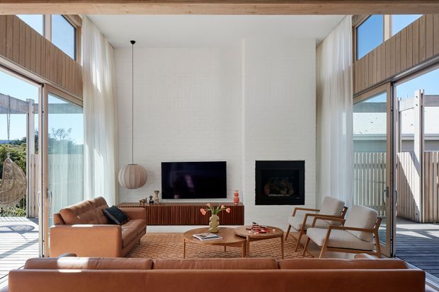 A bright contemporary living room with a high white-painted feature wall, large floor-to-ceiling glazed openings, sheer curtains, and warm timber framing. The room contains tan leather sofas, two light upholstered lounge chairs, a timber slatted media console, television, fireplace, round timber coffee tables, and a woven rug. Outdoor decking and neighbouring structures are visible through the glazing.