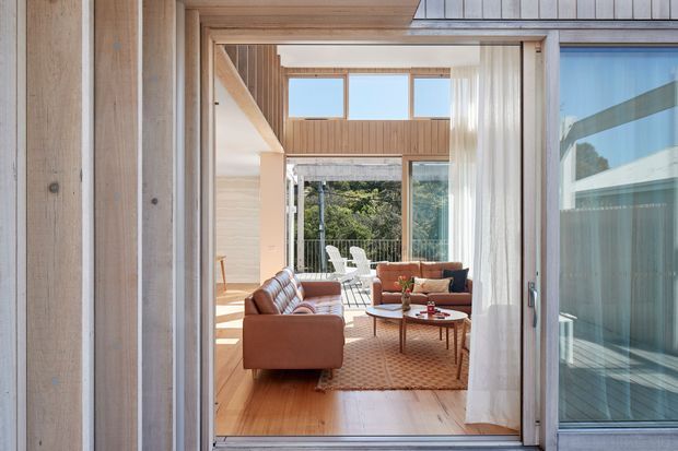 A bright, warm-toned living room viewed through an open glazed doorway. The space features timber-lined walls and ceiling, a high upper-level window band, expansive glass doors, a pair of brown sofas, a patterned rug, and a low wooden coffee table. Outdoor greenery and a patio are visible beyond the glazing.