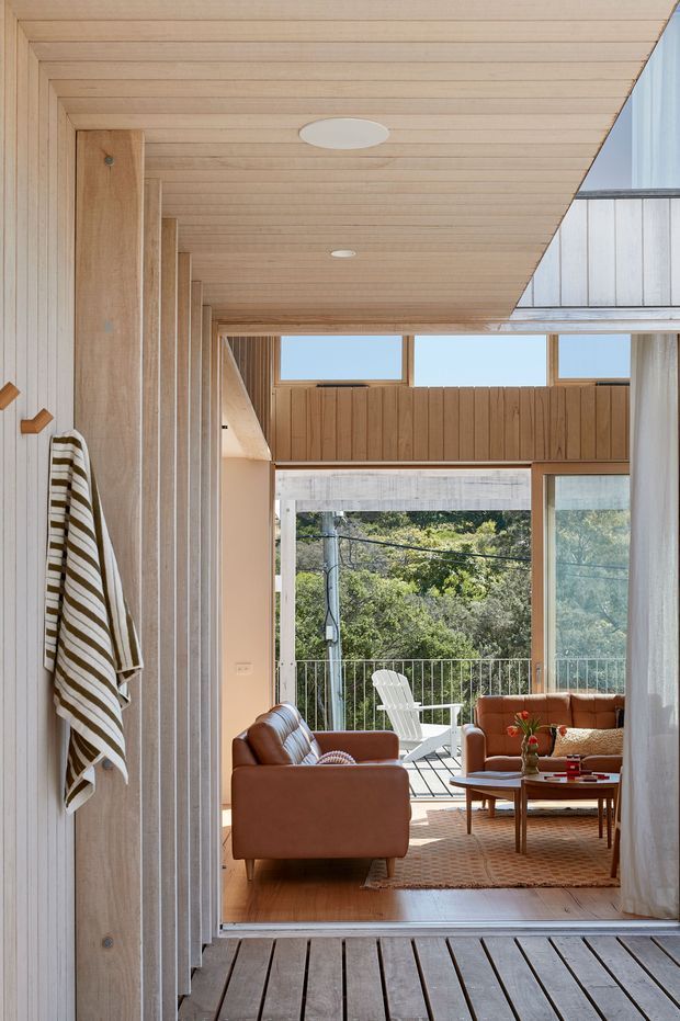 A warm, light-filled residential interior looking from a timber-lined hallway or entry toward a living room and outdoor deck. Vertical and horizontal timber panelling frames large glazed openings, with a timber deck floor, brown leather seating, a low wood coffee table, woven rug, and an outdoor white chair visible beyond. The room has a strong indoor-outdoor connection and high clerestory-style glazing.