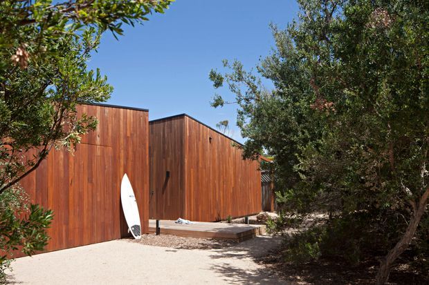 Exterior view of a modern, low-profile building clad in warm vertical timber boards, partially screened by mature landscaping. A small raised timber deck and steps lead to the building, with a surfboard leaning against the wall. The clear blue sky and sandy ground create a relaxed coastal atmosphere.