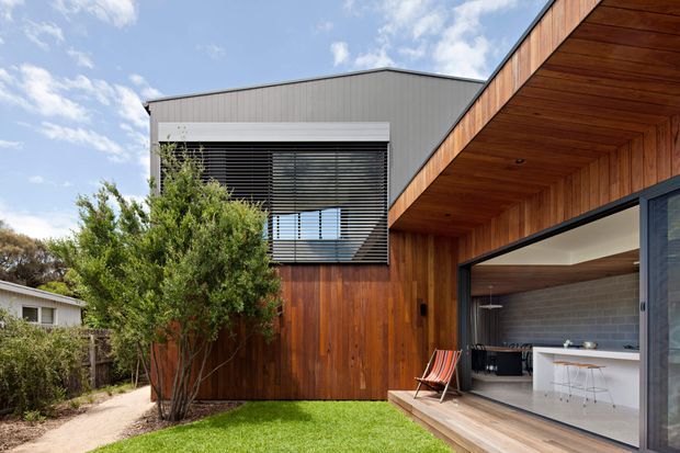 Exterior view of a contemporary two-storey home with a crisp grey upper volume, extensive vertical timber cladding, and a deep timber-lined covered terrace. Large sliding glazing opens to a bright kitchen or entertaining area, while horizontal external blinds shade the upper window. A small landscaped lawn and established planting sit beside the timber deck.