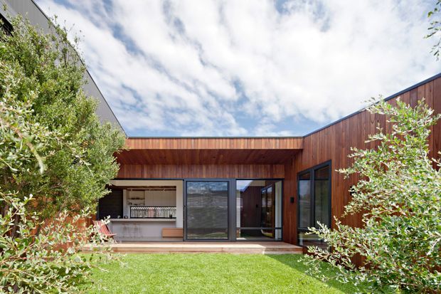 Single-storey contemporary courtyard house viewed from a landscaped lawn, with warm vertical timber cladding, broad glazed openings, a low connecting roofline, and an open-plan interior visible through the central glazing. The U-shaped composition creates a sheltered courtyard with dense planting and a bright sky beyond.