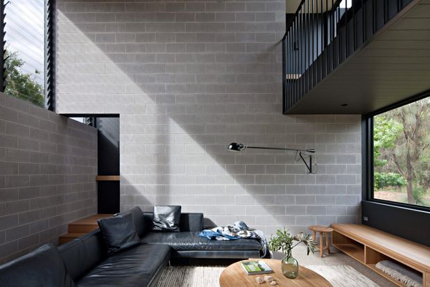 A contemporary double-height living room with exposed grey concrete block walls, a black leather sectional sofa, timber stairs and built-in timber bench seating. Large windows bring in views of surrounding trees, while a black mezzanine-style railing and linear wall light emphasize the vertical architectural volume. A round timber coffee table and sparse greenery add warmth to the otherwise restrained, industrial-inspired interior.