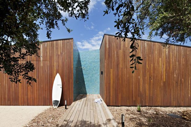 Exterior view of a compact coastal-style structure framed by leafy trees, with tall vertical timber-clad walls, a central turquoise tiled wall, and a timber boardwalk leading toward the entrance. A surfboard leans against the left wall, reinforcing the relaxed beachside setting. Bright natural daylight and warm wood create a casual, inviting atmosphere.