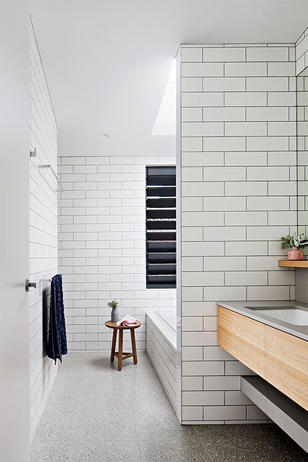 Bright contemporary bathroom with white rectangular subway-tiled walls, a speckled terrazzo-style floor, a skylight, and a black louvred window. The foreground features a timber-front floating vanity with a grey benchtop and integrated basin, while a partially visible bathtub and simple timber stool sit beyond the tiled dividing wall.