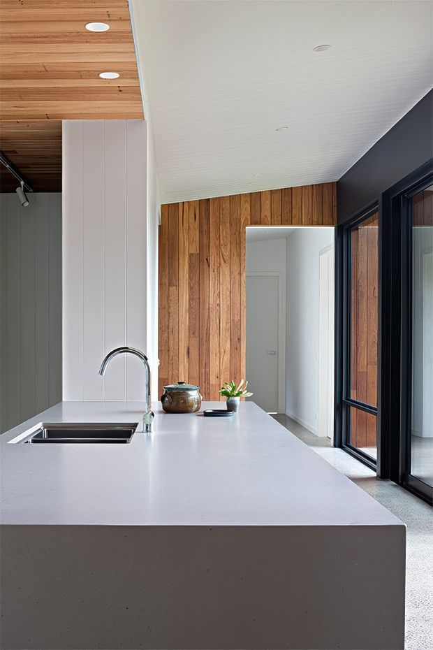 A contemporary kitchen centered on a large white island with an integrated stainless-steel sink and tall curved faucet. Warm vertical timber wall panelling and a timber-lined ceiling contrast with white vertical wall boards, a dark-framed glazed door, and a pale speckled floor. The space is clean and minimally furnished, with a small plant and decorative vessel on the island.