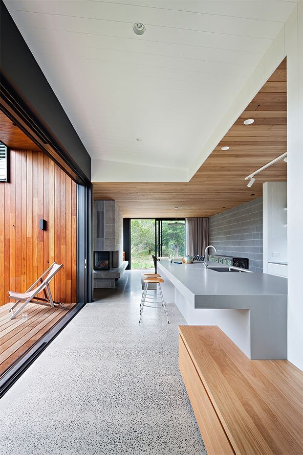 A bright, contemporary open-plan kitchen and living area with a long pale island and integrated seating, dark grey blockwork splashback, extensive warm timber wall and ceiling lining, and a polished aggregate-style floor. Full-height glazing opens to a timber-lined outdoor area on the left, while the rear of the room includes a fireplace and views to greenery. Clean white cabinetry, recessed lighting, and a linear track light create a refined indoor-outdoor composition.