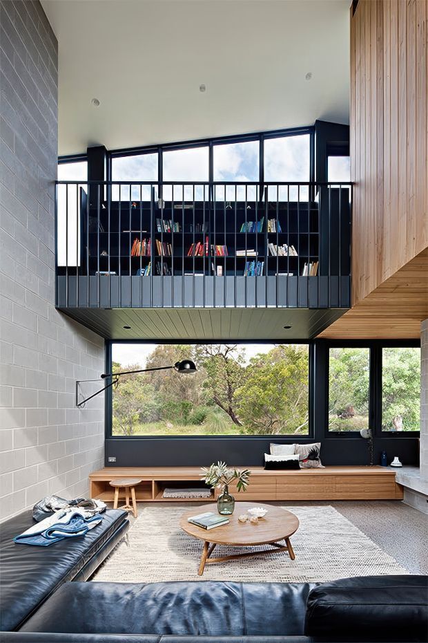 A contemporary double-height living room with a black leather sectional sofa, round timber coffee table, pale textured rug, and built-in timber window seat. A suspended mezzanine library with black metal balustrade and dark shelving overlooks the room. Full-height glazing provides views to greenery, while grey masonry walls, black-framed windows, and a timber-clad upper wall create a warm industrial-modern character.