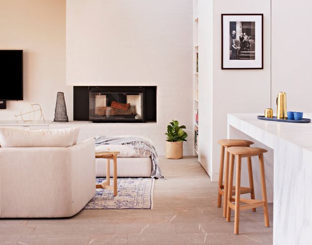 Bright contemporary open-plan living area with a white-painted brick fireplace and inset black-framed firebox, a curved cream sofa, neutral textiles, pale timber-look flooring, and a compact kitchen or bar counter with timber stools. Built-in shelving, framed artwork, indoor greenery, and restrained decor create a warm, refined interior.