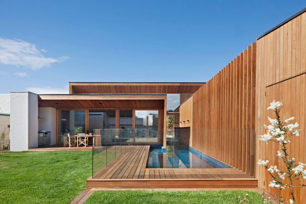 Modern single-storey residential courtyard exterior with a timber-clad privacy fence, flat rooflines, large glazed openings, and a narrow reflecting or swimming pool alongside a timber deck. The composition emphasizes warm timber, crisp white walls, landscaped lawn, and seamless indoor-outdoor connection under a clear sky.