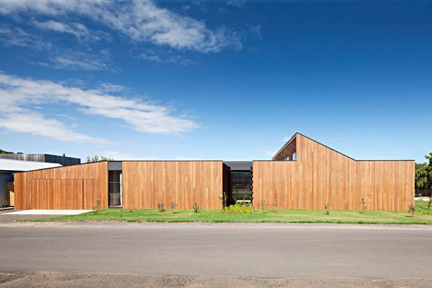 Street-facing view of a low, modern residential building composed of several timber-clad volumes. The facade has warm vertical wood boards, dark recessed glazing and openings, and a prominent pitched gable section rising above the otherwise low roofline. A simple lawn and paved street sit in front under a clear blue sky.