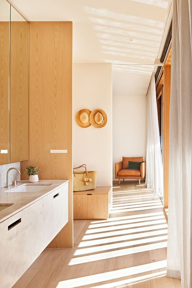 A bright, warm-toned open-plan interior combining a compact kitchen with a long adjoining living or sitting area. The kitchen features full-height light timber cabinetry, a pale stone benchtop with integrated sink, and white lower cabinetry. Strong sunlight filters through sheer curtains, creating pronounced linear shadows across the pale timber floor. A small built-in timber cabinet, woven basket, wall-mounted woven decor, and a caramel leather armchair add natural texture and warmth.