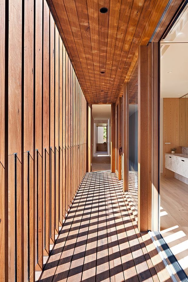A narrow, sunlit timber-lined passageway or covered walkway runs alongside a glazed modern home. The left wall and ceiling are clad in warm vertical and horizontal timber boards, while full-height glazing and timber-framed doors on the right reveal a bright minimalist kitchen with light cabinetry and a stone benchtop. Strong sunlight casts a dramatic grid of shadows across the timber floor, creating a warm, linear architectural composition.