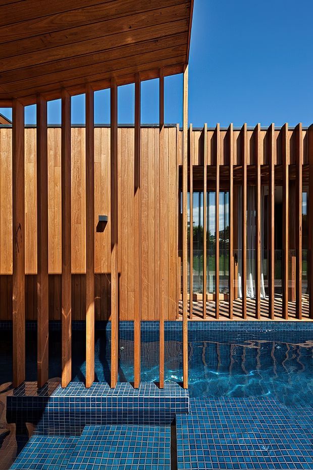 Architectural exterior detail featuring a deep blue tiled pool or water feature in the foreground, surrounded by vertical timber slats and warm timber cladding. Floor-to-ceiling glazing and a timber-lined overhang create a strong connection between the water and the interior, with clear blue sky visible above. The composition emphasizes textured timber screening, reflections, and geometric blue mosaic tiles.