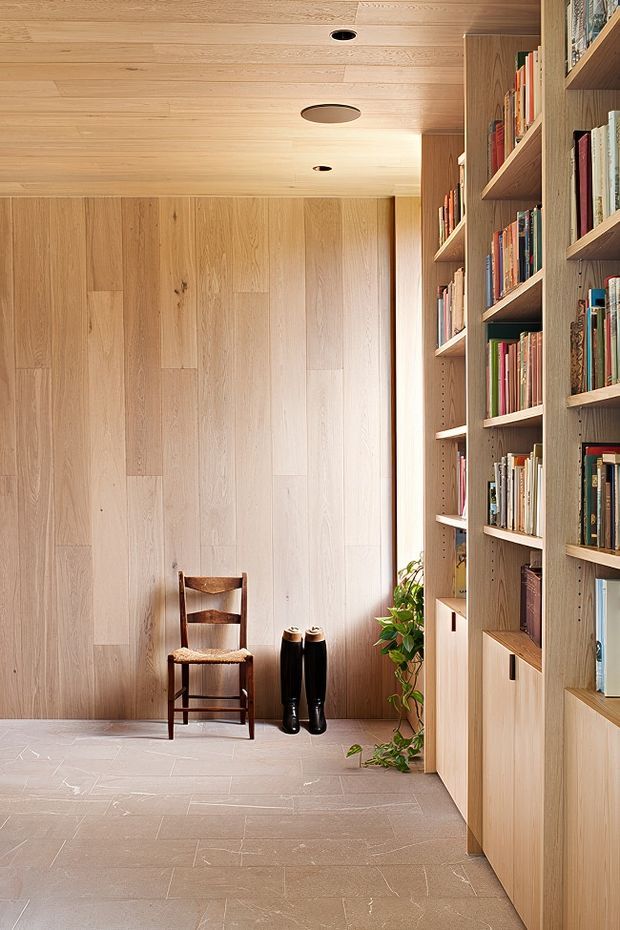 A warm, minimalist entry hall lined with full-height pale timber wall panelling and built-in open bookshelves with lower cabinets. A simple wooden chair, a pair of black boots, and a potted plant sit against the wall. The ceiling is also timber-lined with recessed circular lights, while the floor is finished in large pale stone-look tiles.