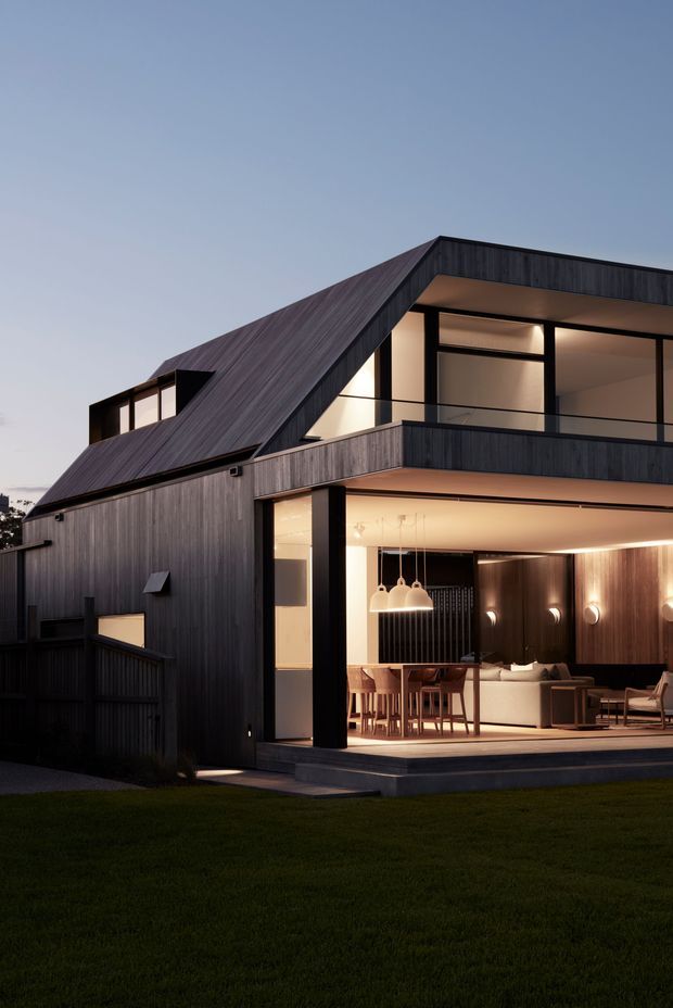 Twilight exterior view of a contemporary multi-level residence with dark vertical timber cladding, a prominent sloping roof, black-framed glazing and a recessed upper balcony. The open ground-floor living and dining area is warmly illuminated and visible through large glazed openings, with a lawn in the foreground.